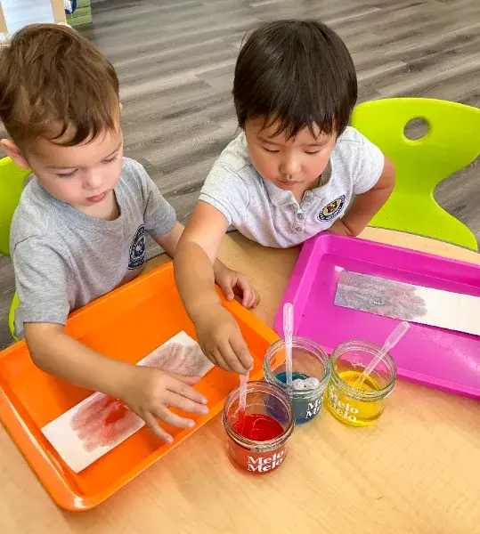 Two children paint with red, blue, and yellow watercolors on paper. They're seated at a wooden table with orange and pink trays.