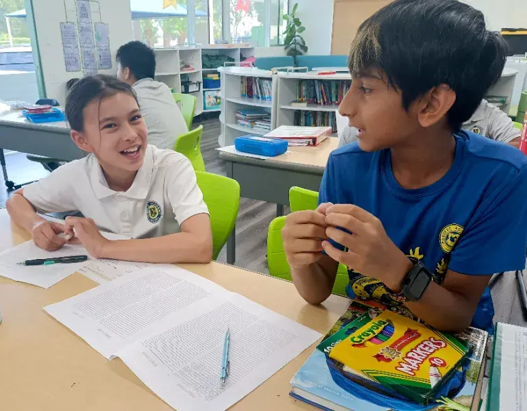 Two students sit at a classroom table, engaged in conversation. Papers and colored markers lie on the table. Bright, cheerful setting.
