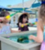 Kids play with toy fishing rods at a water table, smiling under a sunny sky. One girl wears a hat. Background shows a blue building.