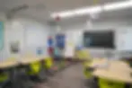 Colorful classroom with yellow chairs, bright posters, and a digital screen. Desks are organized with supplies. Educational charts on walls.