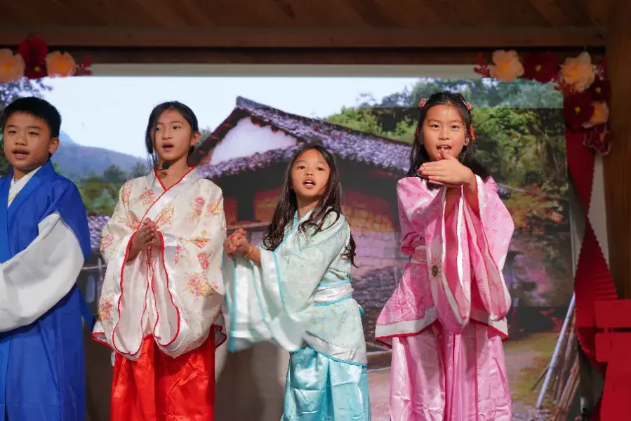 Four children in colorful traditional outfits perform on stage with a scenic backdrop. They appear joyful and expressive.