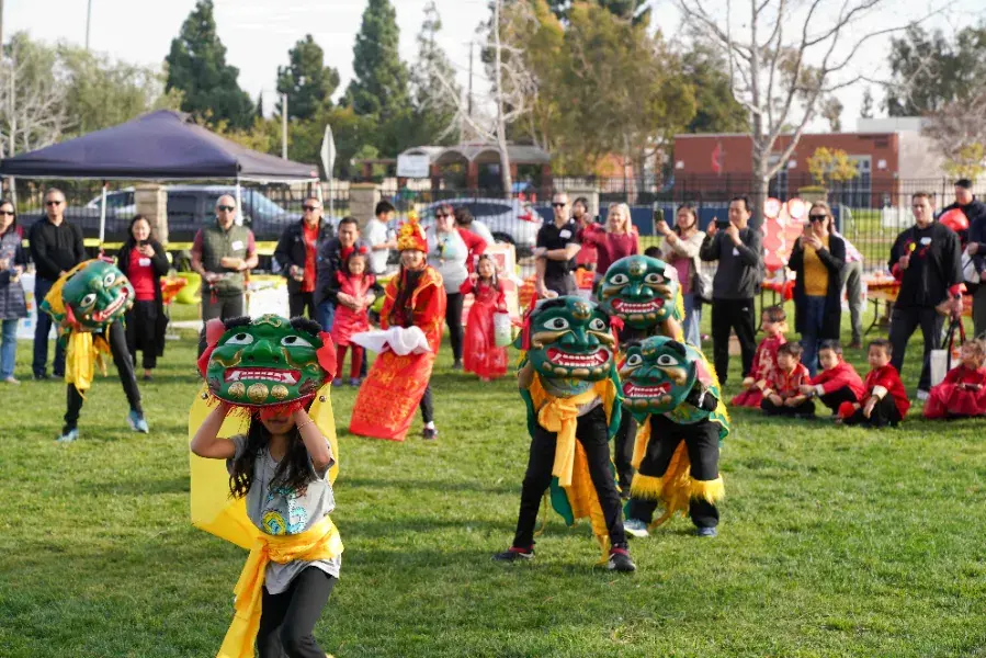 Children perform a dragon dance with green masks and yellow sashes on grass. Onlookers in red attire watch, with trees and a tent in the background.