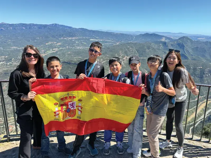 Group of smiling people holding a Spanish flag with a mountainous landscape in the background. Bright, sunny day.