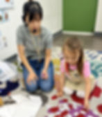 Teacher and child learning with word cards on a colorful rug. The teacher wears a headset. The setting is a classroom.