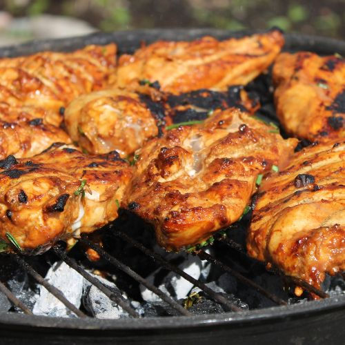 Grilling marinated chicken on a barbecue. Juicy, golden-brown pieces over charcoals, outdoor setting with greenery in the background.