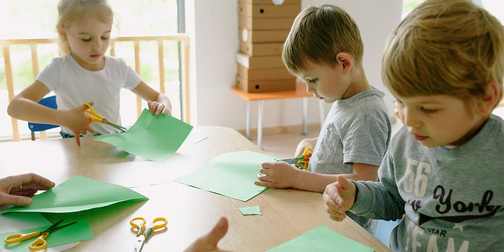 Children cutting green paper with scissors at a table, focused and engaged. Bright room, wooden fence visible outside the window.