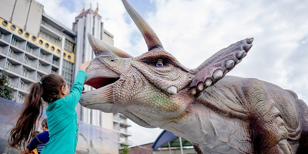 A child in a teal shirt reaches up to touch a triceratops sculpture. Background features a building with circular windows and a cloudy sky.