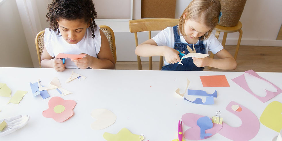 Children focus on cutting colorful paper shapes at a white table in a bright room, with scattered craft materials around them.