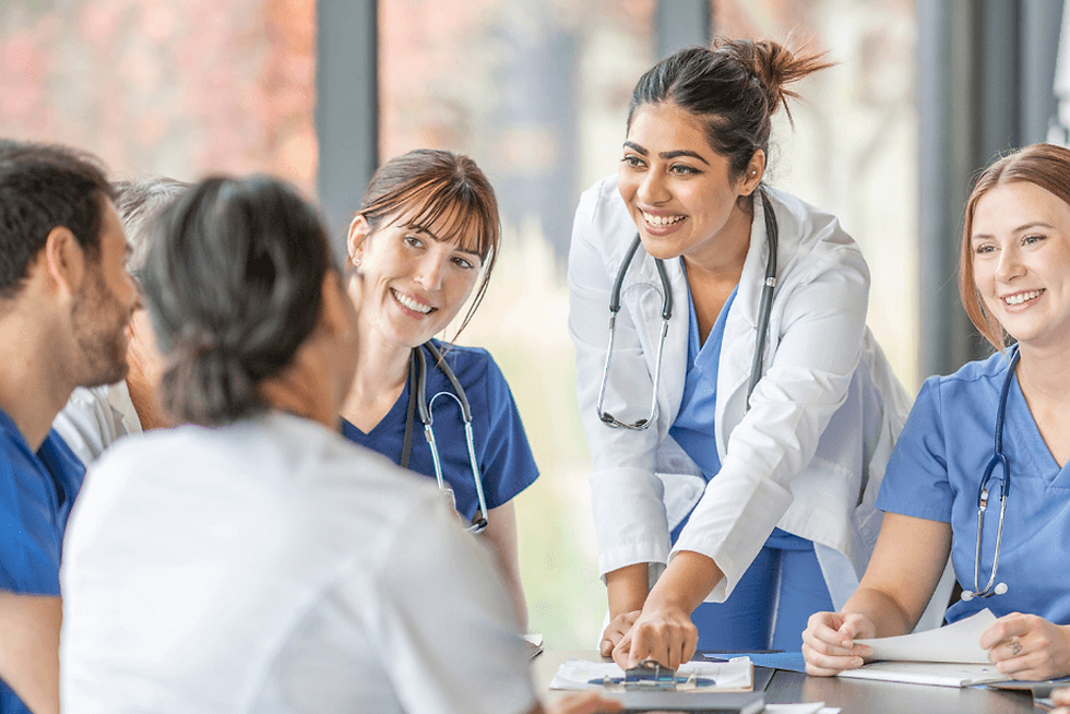 Medical team in blue and white attire, smiling during a meeting in a bright room with large windows. Stethoscopes and papers visible.
