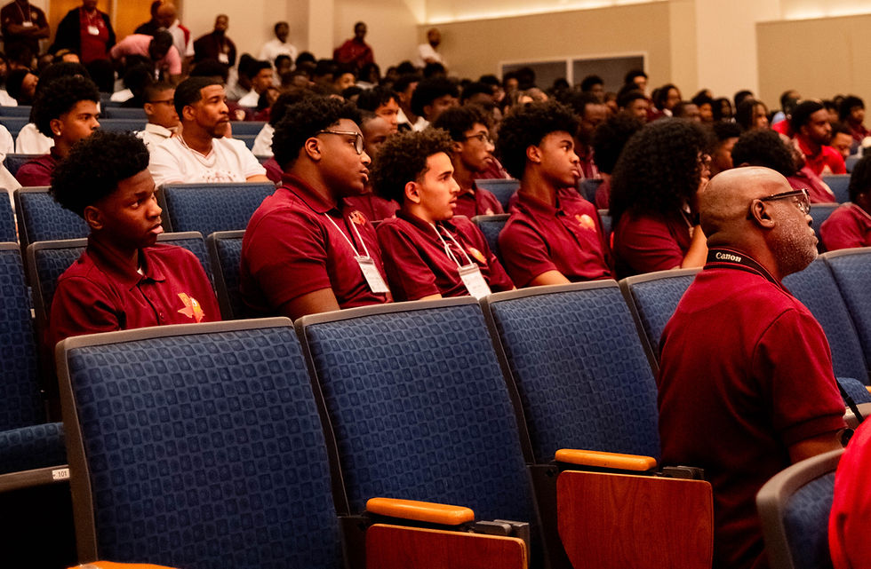 Students wearing maroon shirts sit attentively in an auditorium during a conference session, listening to a speaker while surrounded by a large audience of attendees.
