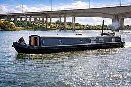 Falcon long black narrowboat cruising along a river beneath a large concrete bridge on a bright sunny day.