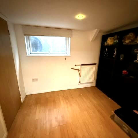 Empty basement room with light wood laminate flooring, small window, white radiator, and a black bookshelf.