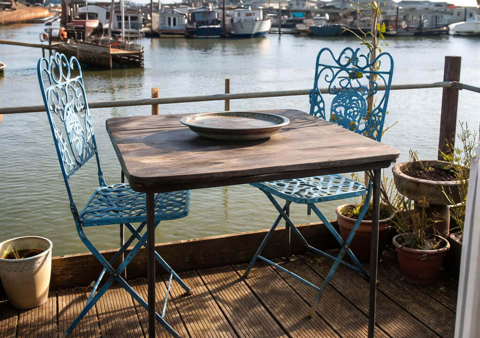 Sunny wooden deck with blue wrought iron table and chairs overlooking boats on a river.