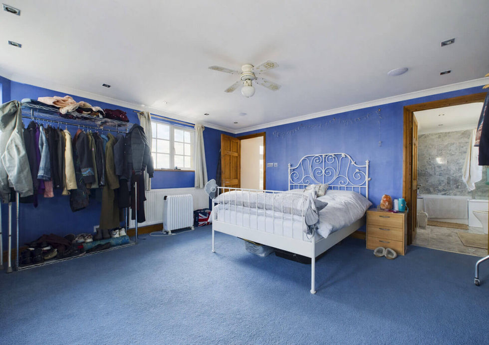 A blue bedroom with a white bed, clothes rack, and open bathroom door. Natural light from windows brightens the cozy space.
