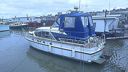 A white motor yacht named 'Atlantis' with a blue canopy docked in a marina with houseboats.
