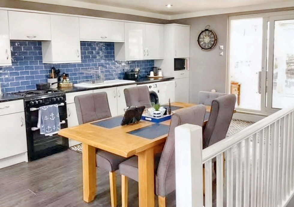 Modern dining area featuring a wooden table surrounded by gray upholstered chairs, with a kitchen and sliding glass doors in the background.