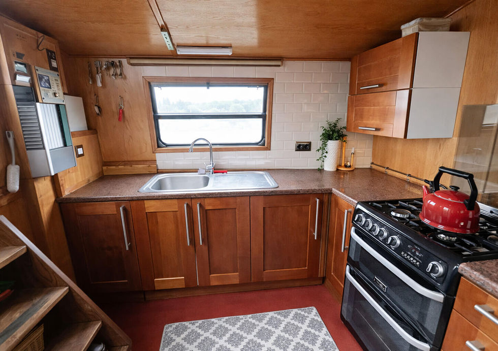 Traditional narrowboat kitchen with wood cabinetry, gas cooker, sink beneath window, and red kettle accent.