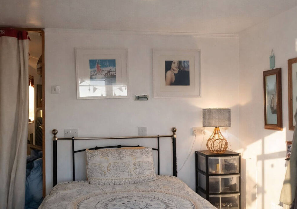 Sunny bedroom with a black and brass bed, patterned duvet, nightstand lamp, and framed art on white walls.