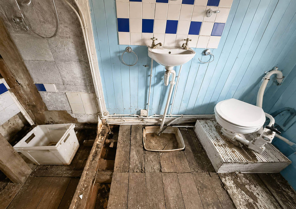 Dilapidated bathroom with blue paneling, damaged wood floor, exposed plumbing, a sink, and a raised toilet.