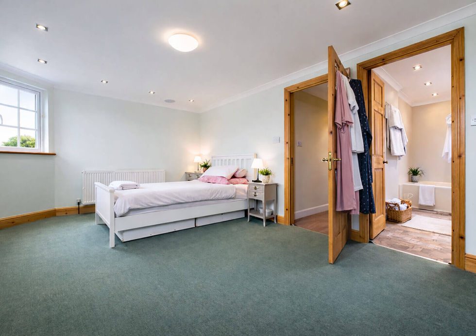 Bright bedroom with a white bed, green carpet, and open door to a bathroom. Clothes hang on the door, and lamps light the room.