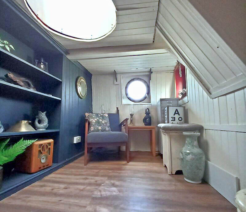 Cozy boat interior with dark blue bookshelf, armchair, wood floor, round window, and rustic decor.