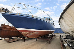Canard boat docked on concrete with other boats and shipping containers in the background.
