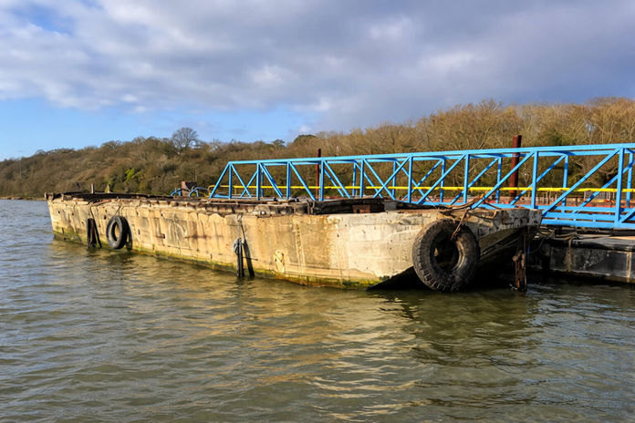 Old concrete barge with a blue bridge sits in the waterway, trees in background.