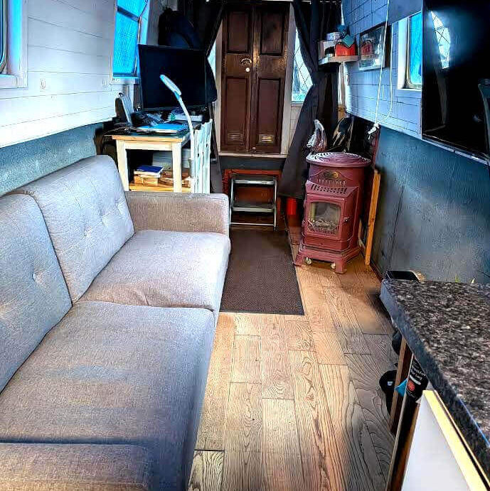 Cozy, narrow interior of a modern tiny home or boat cabin with a gray couch, wood-plank floor, and red stove.