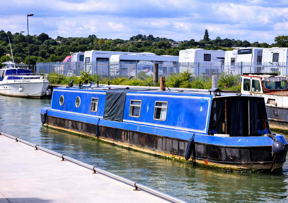 A blue and black narrowboat docked in a canal next to a white speed boat and RV storage area.