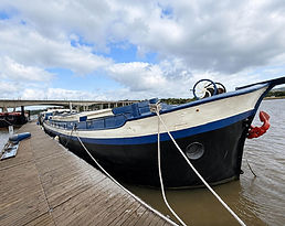 Eersteling blue and black boat docked at a wooden pier with a bridge in the background under a cloudy sky.