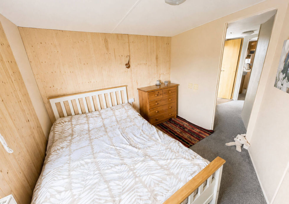 Cozy bedroom with light wood paneling, a bed with a white leaf-patterned duvet, and a wooden dresser.