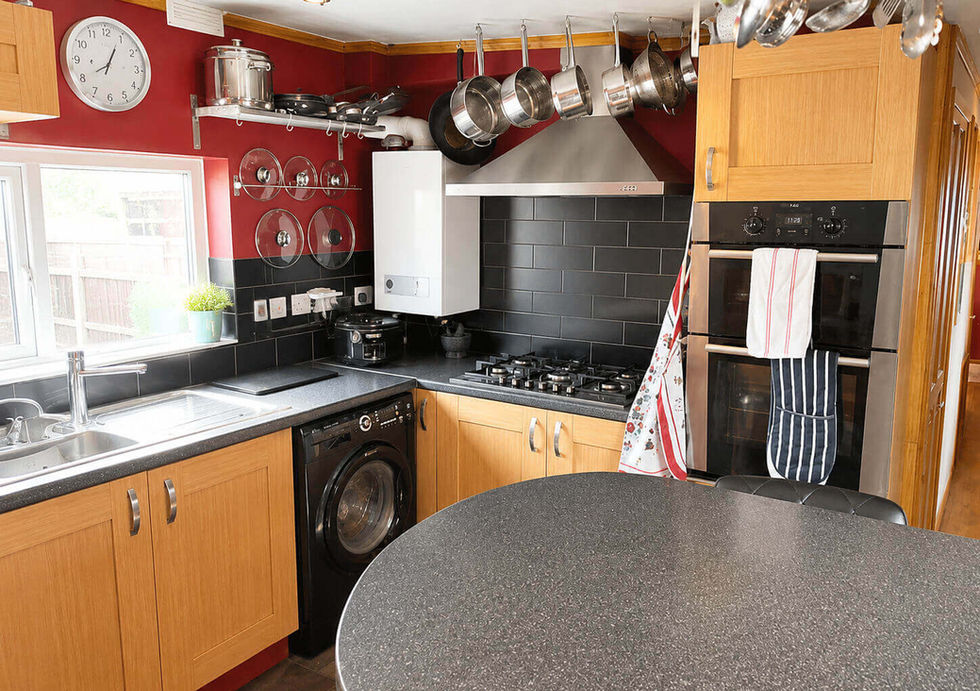 Modern kitchen with red walls, black tile backsplash, light wood cabinets, built-in oven, and a washing machine.