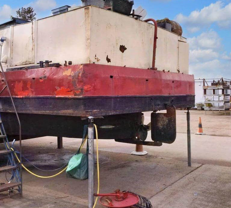 Stern exterior of the boat lifted out of the water on supports, showing the propeller, rudder, and weathered hull.