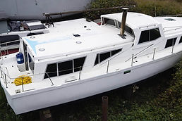 Athos white houseboat docked on land next to another boat, with blue gas can and yellow rope on deck.