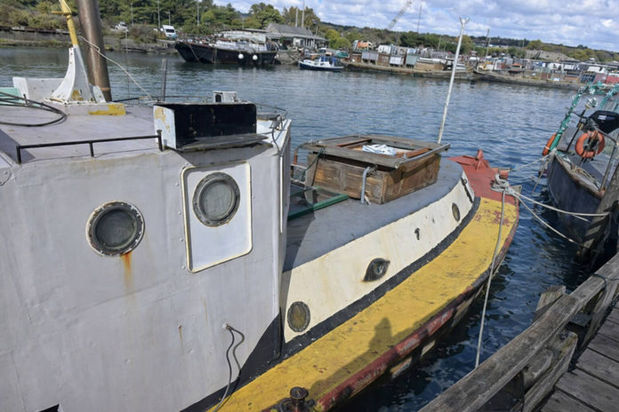 Weathered boat with yellow trim docked in sunny, busy harbor.
