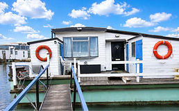 Odean white houseboat with blue accents, a black door, and two orange life rings, docked under a bright blue sky.