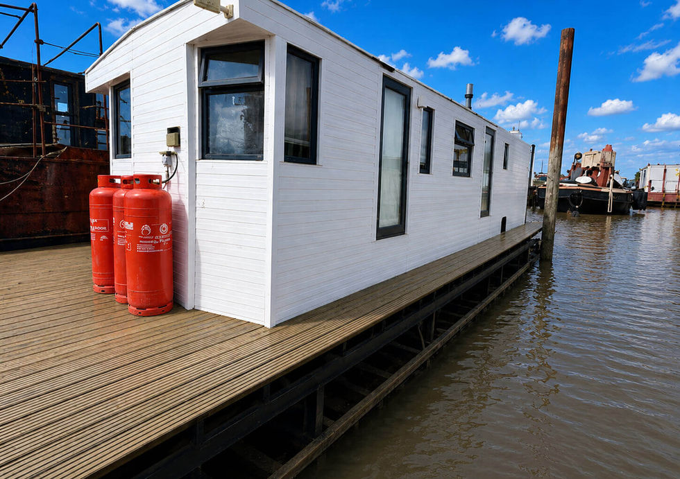 A white modern houseboat with a wooden deck and three red propane tanks, floating on muddy water under a blue sky.