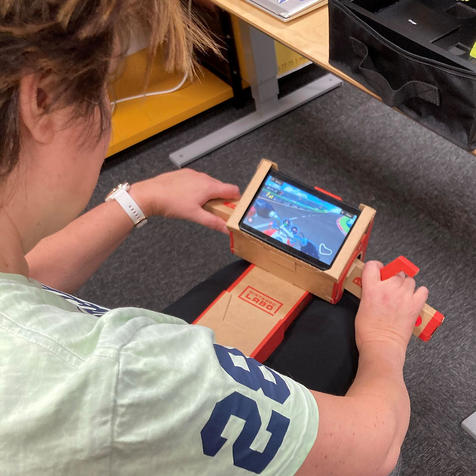 Person with short hair and light green t-shirt holding a cardboard device in their lap resembling motorcycle handlebars. A screen is fastened in the middle, showing a race track.