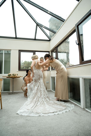 Bridesmaids assisting bride with dress inside a bright room, Cork Wedding, White Amber Photography