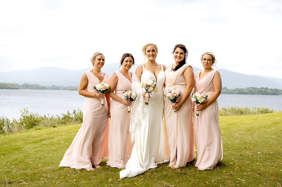 Bride with bridesmaids holding bouquets on a green lawn near lake.