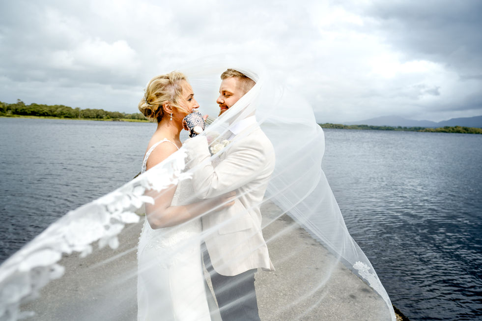 Bride and groom embrace by the water, veil flowing in the wind. White Amber Photography