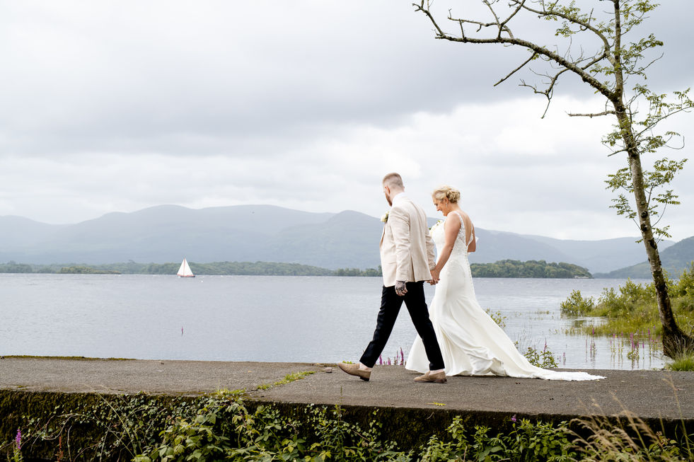 Bride and groom walk near the lake; mountains in background. White Amber Photography – Cork Wedding