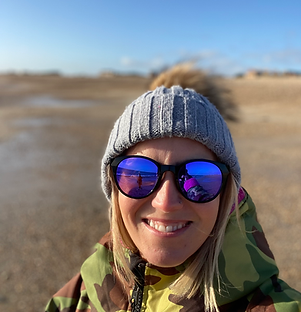A person smiling and looking relaxed at the beach, wearing a dry robe and sunglasses. They stand on the sand with the ocean i