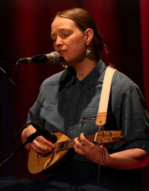 Auf einer schummerigen Bühne sitzt Ulrike im halb Profil vor einem Mikrofon und singt. Sie spielt auf einer Ukulele.
