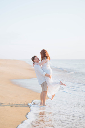 A man in a white shirt and khaki pants lifts a woman in a white dress while waves crash on the beach at their feet.