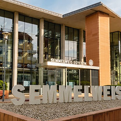 Semmelweis Üniversitesi'nin modern cam cepheli dış mimarisi ve kampüs bahçesinde yer alan büyük beyaz kurum isim tabelası.