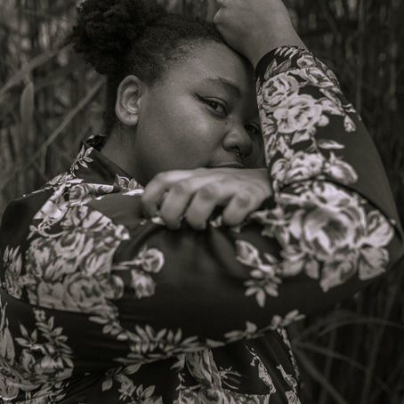 This is a portrait image of Cheniece in black and white. They are wearing a floral long sleeve shirt peering over the right shoulder, focus straight into the camera.