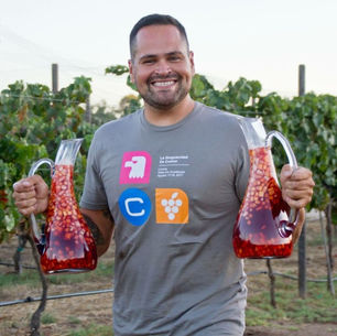 Man standing in a vineyard holding two glass decanters filled with colorful fruit-infused sangria.