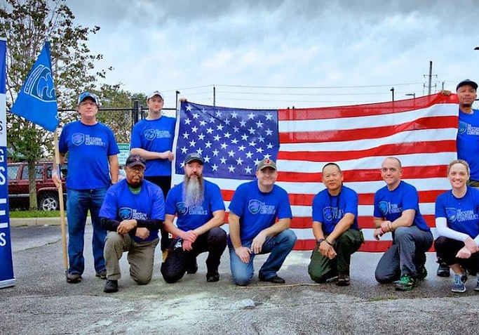The Mission Continues volunteers posing together in front of a large American flag after a community service project.