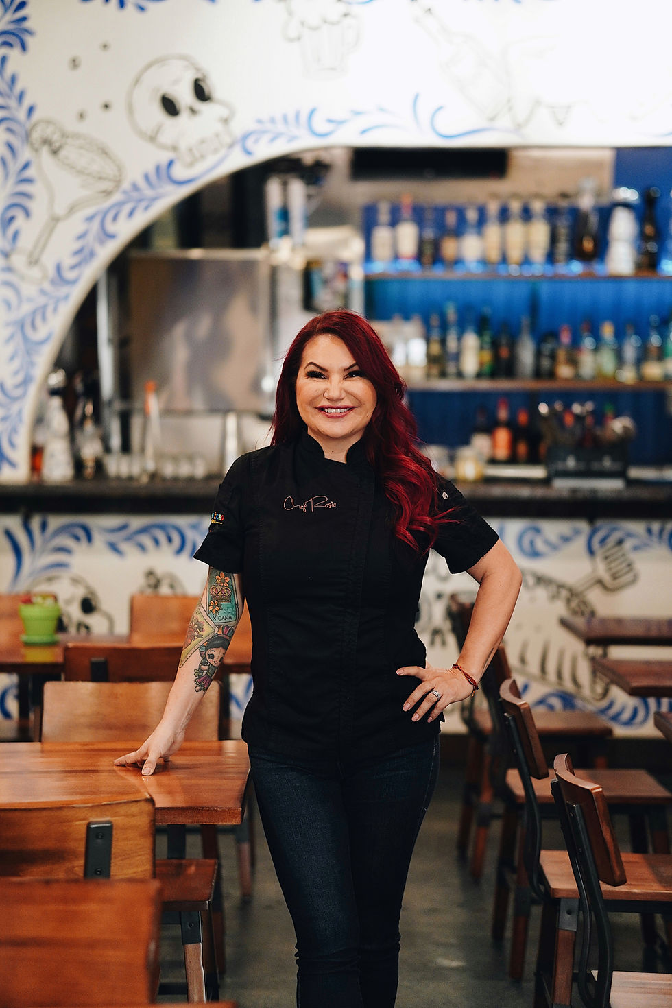 Chef Rosie O’Connor standing in her restaurant dining room wearing a black chef’s coat, with her red hair and colorful arm ta